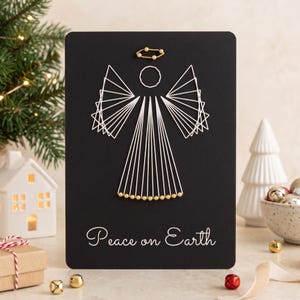 May include: A black rectangular card featuring a white string art angel design. The angel has a circular head, triangular wings, and a dress formed by white thread. A gold bead halo and gold bead hem accent the figure. The text Peace on Earth is printed in white at the bottom.