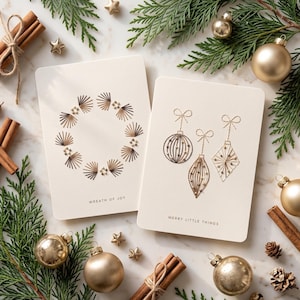 May include: Two off-white rectangular greeting cards. The left card features a gold foil wreath design and the text WREATH OF JOY. The right card displays three gold foil ornaments with bows and the text MERRY LITTLE THINGS.