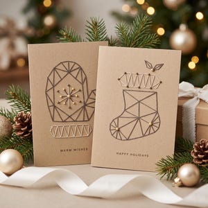 May include: Two tan greeting cards featuring geometric string art designs. The left card displays a mitten with the text WARM WISHES. The right card shows a stocking with the text HAPPY HOLIDAYS. Both cards use dark thread and small pins.