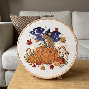 Puede incluir: Un bastidor de bordado circular de madera que contiene un panel de tela con un diseño bordado a mano de un ratón marrón con un sombrero de bruja azul, sentado sobre una gran calabaza naranja rodeada de calabazas más pequeñas y hojas.