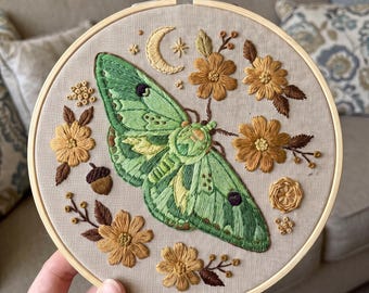 May include: A circular wooden embroidery hoop containing a beige fabric panel with a hand-stitched design. The central feature is a large green moth with dark purple spots, surrounded by golden-yellow flowers, brown leaves, an acorn, and a crescent moon.