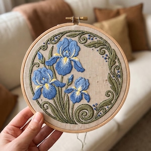 May include: A circular wooden embroidery hoop containing a piece of beige fabric with hand-stitched blue iris flowers, yellow centres, and green leaves. The floral design is framed by an intricate, swirling green vine border with small blue accents.