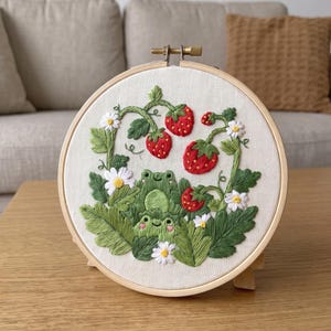 May include: A circular wooden embroidery hoop containing a fabric panel with hand-stitched thread art. The design features two green frogs nestled among lush green leaves, white daisy flowers, and bright red strawberries with yellow seeds.