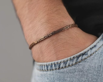 Puede incluir: Una pulsera fina y flexible compuesta por numerosas cuentas diminutas de color bronce metálico dispuestas en un patrón cilíndrico denso. La pulsera presenta una superficie uniforme y texturizada con un brillo sutil alrededor de la muñeca.