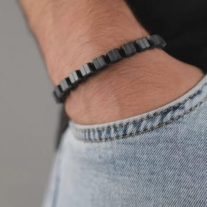 May include: A bracelet composed of a series of matte black, square-shaped beads linked together in a continuous row. The beads feature a smooth, uniform finish and are arranged to form a flexible band that wraps around the wrist area.