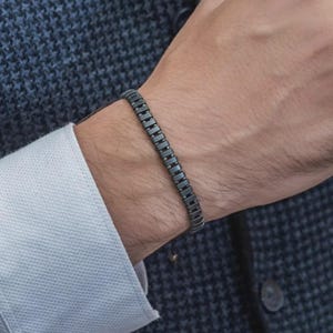 May include: A dark gray bracelet composed of small, rectangular, metallic links arranged in a continuous, uniform row. The bracelet features a smooth, matte finish and sits snugly around the wrist, showcasing a structured and repetitive geometric design.