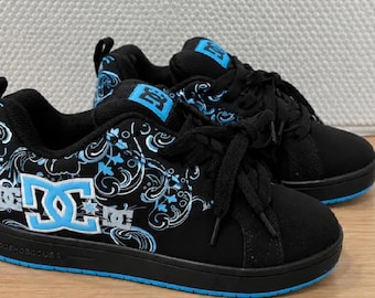 May include: Black DC sneakers featuring a bright blue paisley-inspired graphic pattern on the side panels. The shoes have black laces, a padded tongue with a blue DC logo patch, and a solid black sole with a thin blue stripe along the bottom.