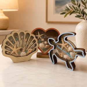 May include: Three wooden shadow box displays in the shapes of a seashell, a heart, and a sea turtle. Each frame contains sand, small seashells, and starfish. The seashell frame reads "EVERY SEASHELL TELLS A STORY" and the turtle reads "Tommy and Marilyo".