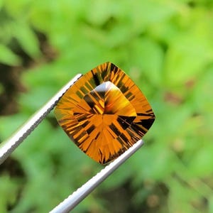 May include: A cushion-cut citrine gemstone with a deep golden-orange hue and intricate, radiating geometric facets. The transparent stone displays sharp, symmetrical light reflections across its surface, held by a pair of silver-toned metal tweezers.
