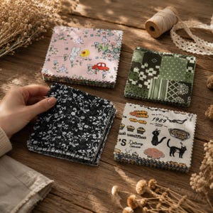 May include: Four stacks of square fabric swatches featuring diverse patterns including a pink background with a white rabbit and red car, a green geometric print, a black floral design, and a cream print with cats and the text Kiki's 1989.