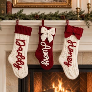 May include: Three cable-knit Christmas stockings hanging in a row. The left stocking is cream with a dark red cuff and the name Daddy in red. The centre is dark red with a cream bow and the name Avery in cream. The right is cream with a red bow and the name Honey in red.