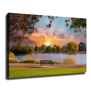 May include: A rectangular canvas print featuring a landscape scene of a black metal park bench positioned on a grassy bank overlooking a calm lake. The background displays a vibrant sunset with orange and pink clouds, framed by tree branches.