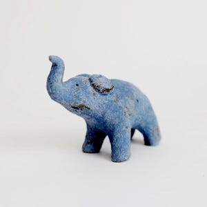 May include: A small, hand-sculpted elephant figurine with a textured, matte blue finish. The elephant is depicted in a standing position with its trunk raised upward, featuring subtle dark grey speckling across its surface and rounded ears.