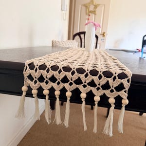 May include: A beige crochet table runner featuring an open diamond pattern. The edges are finished with a row of light-colored wooden beads and matching fabric tassels that hang down from the ends of the runner, creating a decorative border.