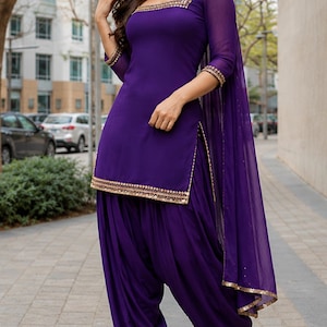 Könnte beinhalten: Ein tiefviolettes Salwar-Kameez-Set bestehend aus einer knielangen Tunika mit Dreiviertelärmeln und quadratischem Ausschnitt, kombiniert mit einer plissierten Salwar-Hose und einem passenden transparenten Dupatta. Die Ränder sind mit Gold verziert.