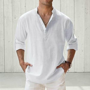 May include: A white long-sleeve shirt with a mandarin collar and a partial button-down placket. The shirt features a textured fabric, cuffed sleeves, and a straight hemline. It is worn with a silver chain necklace and a brown leather wristband.