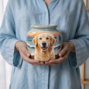 Op de afbeelding: Een keramische pot met deksel voorzien van een geschilderde illustratie van een golden retriever op een strand met oceaangolven, een zonsondergang en een surfplank. De pot heeft een gladde, ronde vorm en een glanzende afwerking met gedetailleerd kunstwerk.