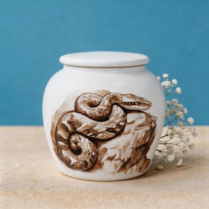 May include: A white ceramic jar with a lid, featuring a detailed brown and tan painted illustration of a coiled snake resting on a rock. The jar has a smooth, rounded body and a matching removable lid, showcasing a realistic wildlife art design.