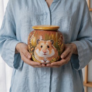Könnte beinhalten: Ein rundes Keramikgefäß mit Deckel, das eine handgemalte Illustration eines braun-weißen Hamsters zeigt, umgeben von goldenen Weizenähren und Blattwerk. Das Kunstwerk hat eine texturierte Pinselstrich-Optik in warmen Orange- und Brauntönen.