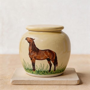 Puede incluir: Un tarro de cerámica color crema con tapa a juego, que presenta un caballo marrón pintado a mano sobre hierba verde. El caballo está representado de perfil, mirando hacia la izquierda con la cabeza levantada, sobre un acabado liso.