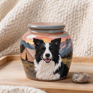 Op de afbeelding: Een keramische urn met een geschilderde scène van een zwart-witte Border Collie hond voor een kampeertent en een berglandschap bij zonsondergang. De urn heeft een afgerond lichaam en een bijpassend deksel met een gladde, glanzende afwerking.