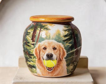 Puede incluir: Una urna de cerámica con la pintura de un golden retriever que sostiene una pelota de tenis de color amarillo brillante en la boca. El fondo representa un bosque verde y frondoso con árboles altos. El recipiente tiene una tapa de madera redondeada.