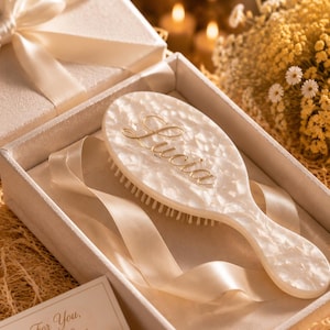 May include: A white pearlescent hairbrush with the name Lucia written in gold script, resting inside a cream-coloured gift box with a satin ribbon. A small rectangular card with the text For You, With Love is placed in the corner of the box.