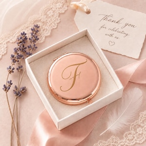 Può includere: Uno specchietto da borsa rotondo in oro rosa con una lettera F in corsivo incisa sul coperchio, presentato in una scatola regalo quadrata bianca. La scatola include un cartellino con il testo Thank you for celebrating with us e un piccolo cuore.