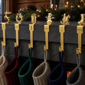 May include: Six gold-toned metal stocking holders with personalized names engraved on the vertical plates. The toppers feature a reindeer, wreath, gift box, snowman, sleigh, and boot. Each holder supports a knit stocking hanging from a hook.