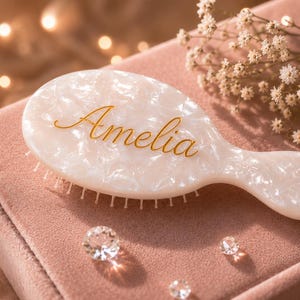 May include: An oval hairbrush with a pearlescent white finish and a marbled texture. The name Amelia is written in gold cursive script across the center of the brush head. The handle is short and integrated into the main body of the brush.