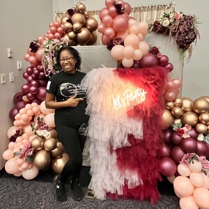 May include: A woman stands in front of a backdrop decorated with balloons in shades of pink, burgundy, and gold. The balloons are arranged in a large arch shape. The backdrop also features a white and burgundy tulle curtain with a neon sign that reads "Let's Party".