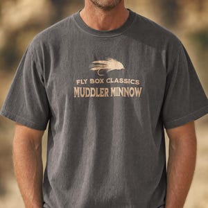 May include: A charcoal grey short-sleeve t-shirt featuring a tan graphic of a fishing fly. The text above the fly reads FLY BOX CLASSICS, and the text below the fly reads MUDDLER MINNOW. The shirt has a crew neckline and a standard fit.