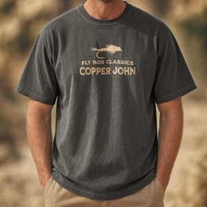 Könnte beinhalten: Ein anthrazitfarbenes Kurzarm-T-Shirt mit einer beigen Grafik einer Angel-Fliege und dem Text FLY BOX CLASSICS COPPER JOHN auf der Brust. Das T-Shirt hat einen Rundhalsausschnitt und eine lockere Passform mit einer verwaschenen Stoffstruktur.