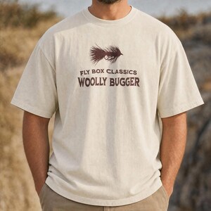 Può includere: Una t-shirt a maniche corte color crema con una grafica marrone scuro raffigurante una mosca da pesca. Il testo stampato in marrone scuro recita FLY BOX CLASSICS e WOOLLY BUGGER. La maglietta presenta un girocollo e una vestibilità standard.