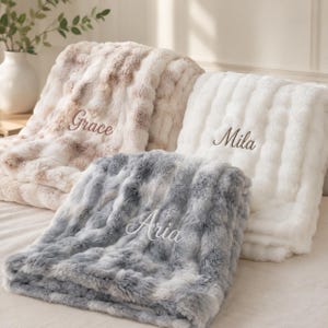 Può includere: Tre coperte in pelliccia sintetica testurizzata nei colori bianco, grigio e una miscela beige-bianco. Ogni coperta presenta un nome ricamato in corsivo: Grace sulla coperta beige, Mila su quella bianca e Aria su quella grigia.
