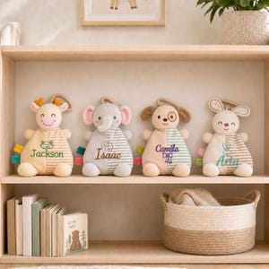 May include: Four plush animal security blankets featuring a deer, elephant, dog, and rabbit. Each toy has a split-colour design with one side solid and one side striped, embroidered with names Jackson, Isaac, Camila, and Aria, and small icons.