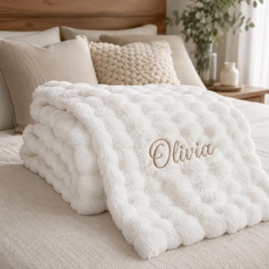 May include: A white plush blanket with a textured bubble pattern and the name Olivia embroidered in a light brown cursive script. The blanket is folded and features a soft, fuzzy surface with a repeating circular design across the fabric.