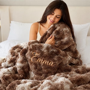 May include: A plush, textured faux fur throw blanket in shades of brown and cream. The blanket features the name Emma embroidered in a cursive script font using a light brown thread. The material has a thick, shaggy pile with a soft appearance.