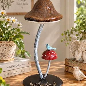 Könnte beinhalten: Eine dekorative Metallskulptur mit zwei Pilzen auf einem runden schwarzen Sockel. Der größere Pilz hat einen strukturierten braunen Hut, während auf dem kleineren roten Pilzhut ein kleiner blauer Vogel sitzt. Der Name Amelia ist in Gold gedruckt.