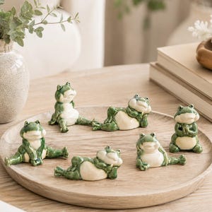 May include: A set of five small ceramic frog figurines in various poses, including sitting, reclining, and praying. Each frog features a glossy green finish on the back and head with a smooth cream-coloured belly, arranged on a round wooden tray.