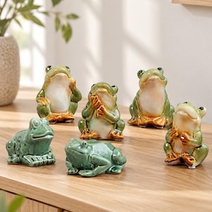 Może przedstawiać: Kolekcja sześciu ceramicznych figurek żab w różnych pozach. Trzy żaby siedzą wyprostowane z jasnymi brzuchami i zielonymi grzbietami, podczas gdy trzy pozostałe przedstawiono w różnych pozycjach spoczynkowych z jednolitą zieloną glazurą.