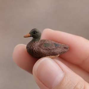 Może przedstawiać: Miniaturowa figurka kaczki o ciemnobrązowym korpusie, teksturowanej powierzchni, ciemniejszej głowie i jasnobrązowym dziobie. Mała kaczka jest wyrzeźbiona z widocznymi detalami piór i małą niebieską plamką na skrzydle, w kompaktowym rozmiarze.