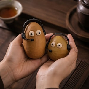 Op de afbeelding: Twee gladde, bruine, ovaalvormige objecten met cartoonachtige witte ogen en zwarte pupillen. Elk object draagt een kleine zwarte headset met een microfoon aan de zijkant. De objecten worden samen vastgehouden, waarbij de ene duidelijk groter is.