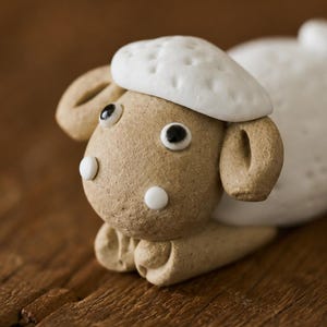 Puede incluir: Una pequeña figura de arcilla de una oveja con cabeza y cuerpo de color tostado, que presenta un gorro de lana blanca texturizada en la cabeza, dos pequeños puntos circulares blancos en la cara y dos ojos de color blanco y negro.