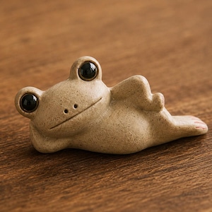 Może przedstawiać: Mała, beżowa, nakrapiana ceramiczna figurka żaby w pozycji leżącej. Żaba ma duże, błyszczące, czarne oczy, prostą poziomą linię zamiast ust i dwa małe nozdrza. Ciało figurki ma fakturę z drobnymi, ciemnobrązowymi kropkami na całej powierzchni.