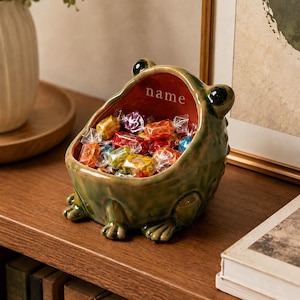 Op de afbeelding: Een groene keramische kom in de vorm van een kikker met een gladde, glanzende afwerking en een wijde, open mond. De binnenkant van de kom is diep rood-oranje van kleur en bevat het woord name in witte letters. De kom is gevuld met snoepjes.