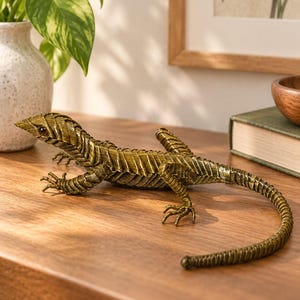 Puede incluir: Una figura decorativa de lagarto hecha de alambre metálico enrollado en un tono verde oliva apagado. La escultura presenta un cuerpo texturizado y segmentado, cuatro patas extendidas con garras distintas, una cola larga y delgada y ojos oscuros como cuentas.