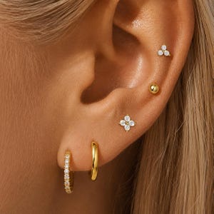 May include: A set of gold-toned earrings consisting of two small hoop earrings, one plain and one with clear stones, a four-stone flower stud, a plain gold ball stud, and a three-stone triangular stud, all worn in multiple ear piercings.