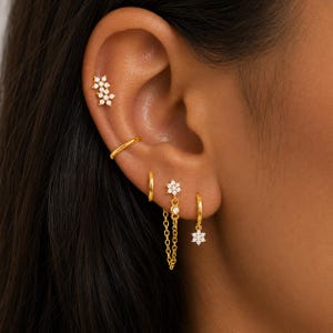 May include: A set of gold-toned earrings featuring a double-flower stud, a plain hoop, a flower stud connected to a hoop by a delicate chain, and a single hoop with a flower charm, all accented with small clear stones set in a polished metal finish.