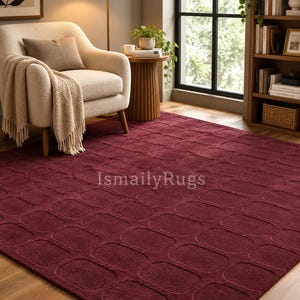 May include: A deep burgundy area rug featuring a repeating pattern of raised, rounded rectangular shapes across its surface. The rug displays the text IsmailyRugs in the center. The texture appears soft and plush, covering the floor area.